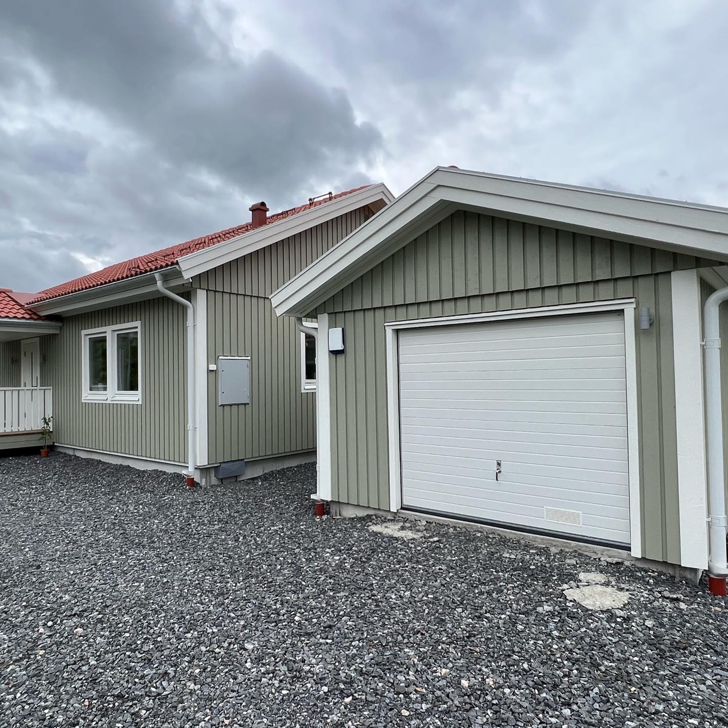 Grönmålat trähus och garage med vit garageport, vita knutar och vita fönster mot en grusplan