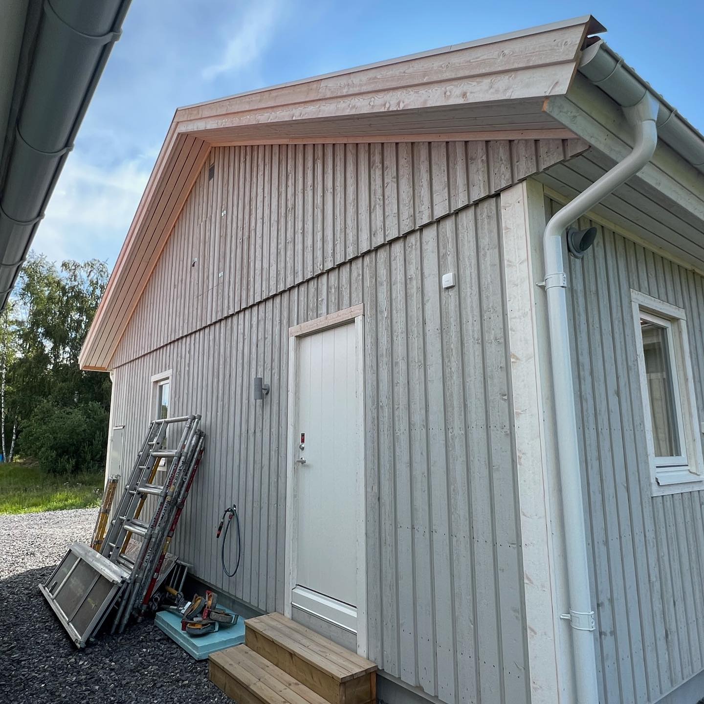 Grå träfasad före målning med stegar och ställningsdelar lutade mot väggen intill en vit dörr