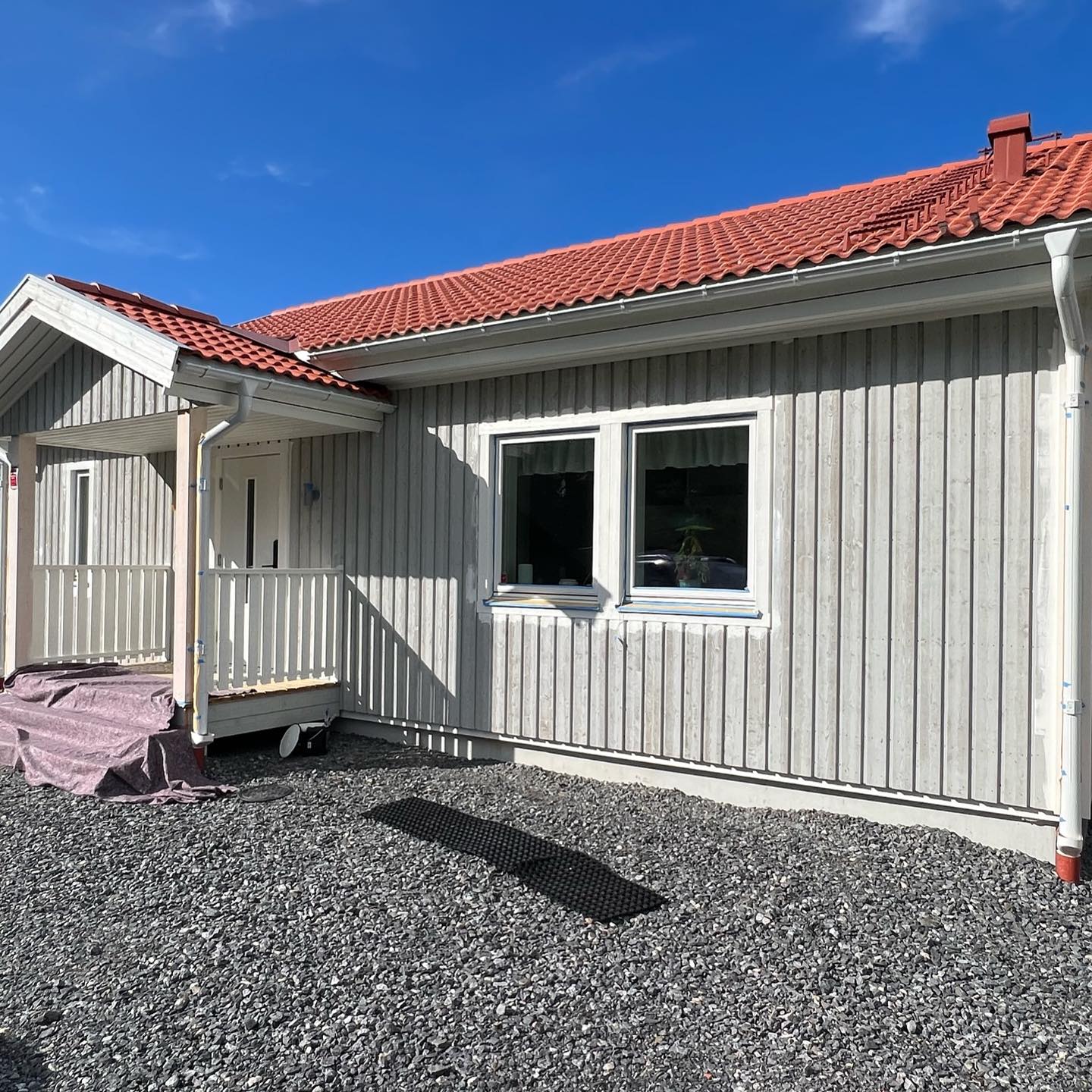 Långsida på grått trähus före målning med vita fönster och skyddstäckt entré under skärmtaket