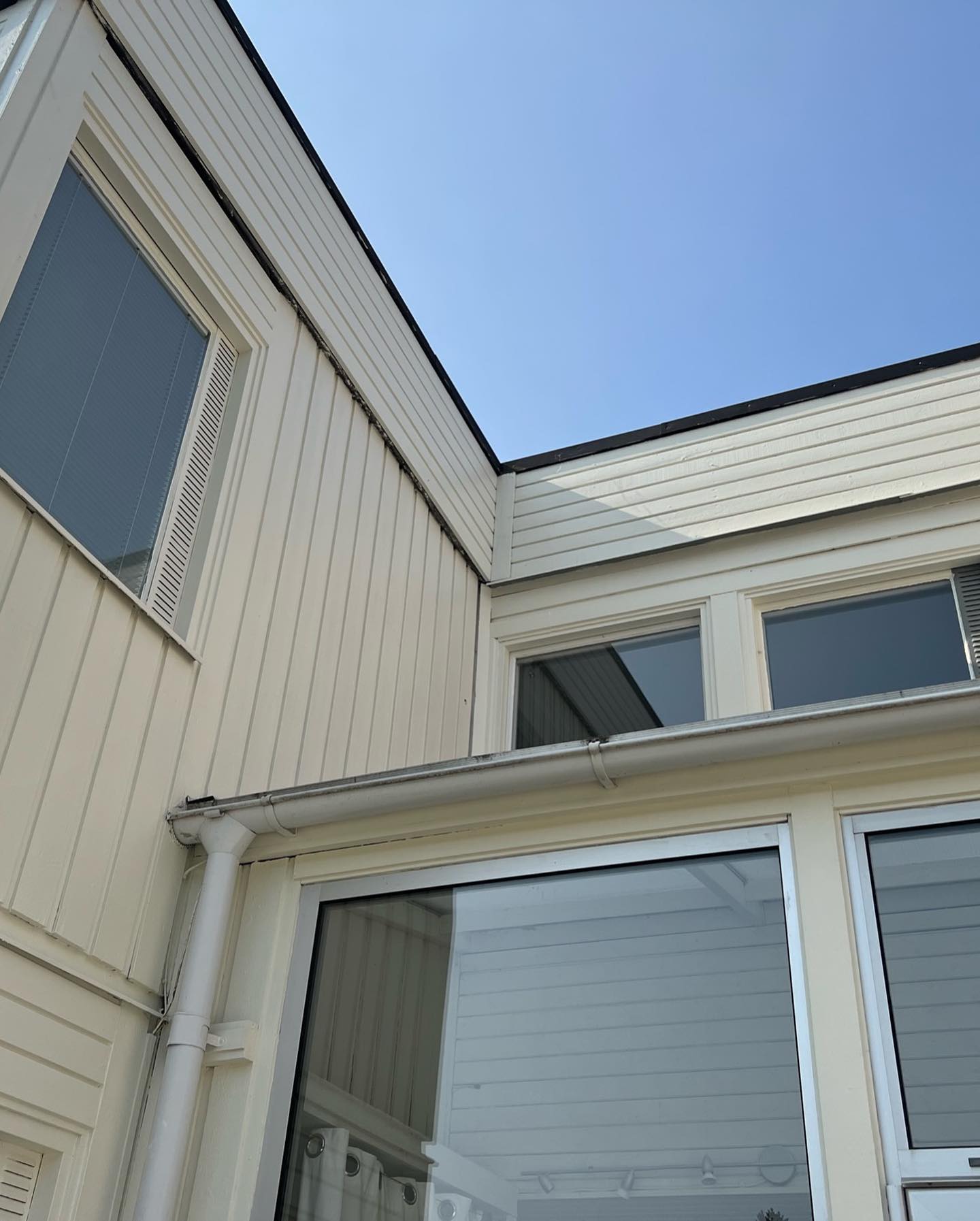 Ljus träfasad med stående panel, vita fönster och stuprör runt ett glasat entréparti