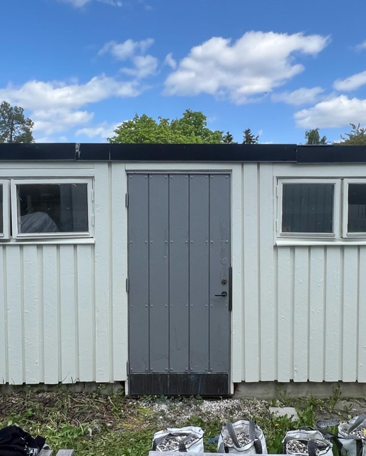 Nymålat förråd med ljus stående panel, vita fönster och grå dörr med plåtsockel