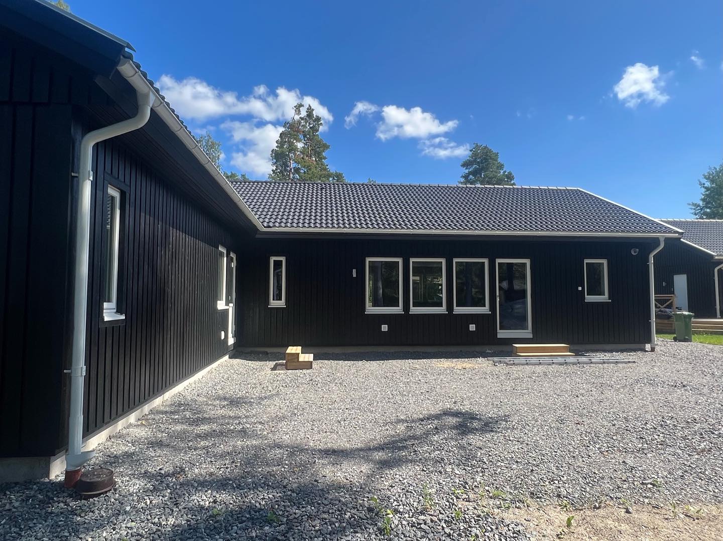 Vinkelbyggd enplansvilla med svart stående träpanel, vita fönster och vita stuprör efter målning