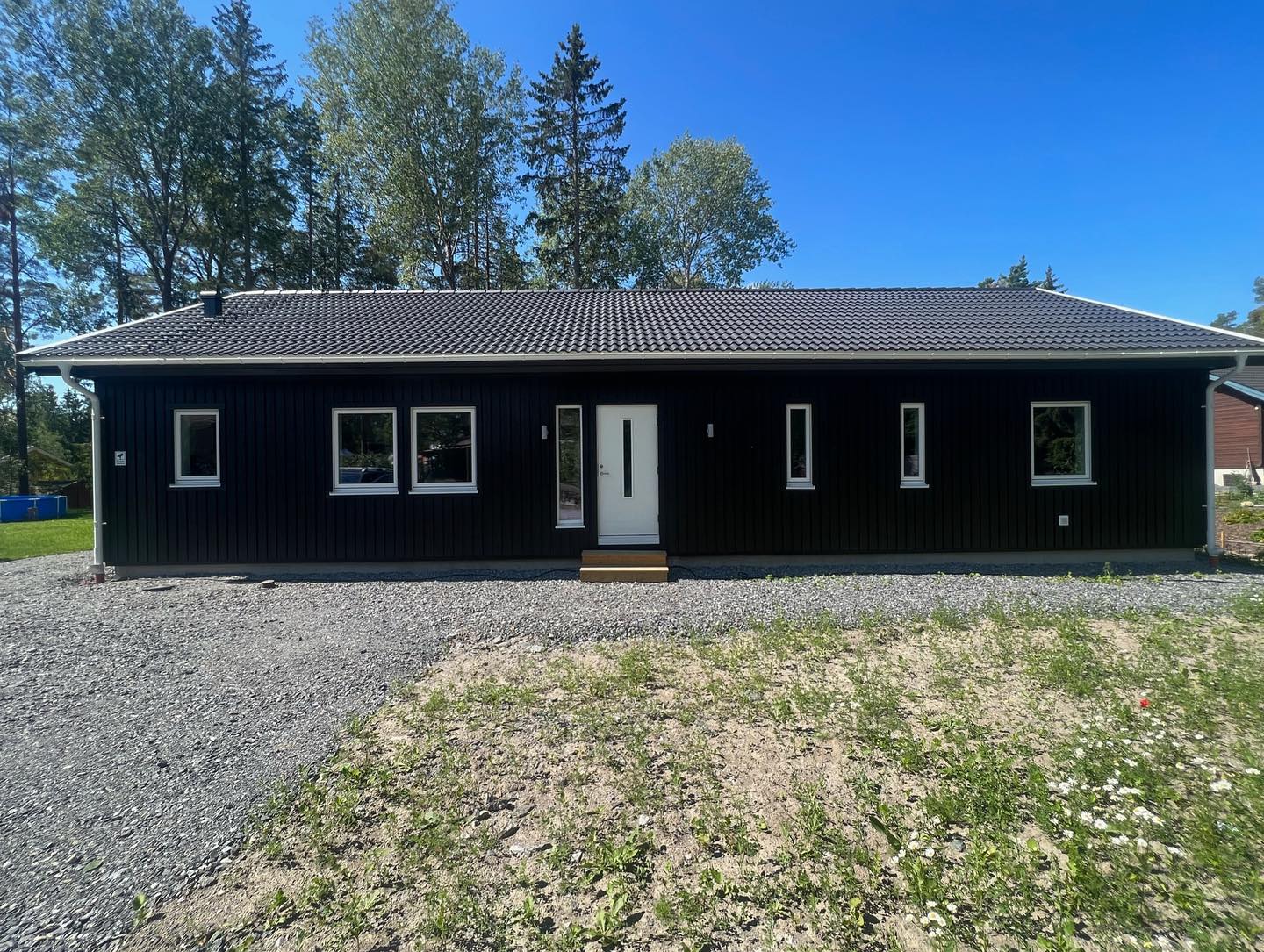 Svart enplansvilla framifrån med vit entrédörr, vita fönster och stående träpanel efter målning